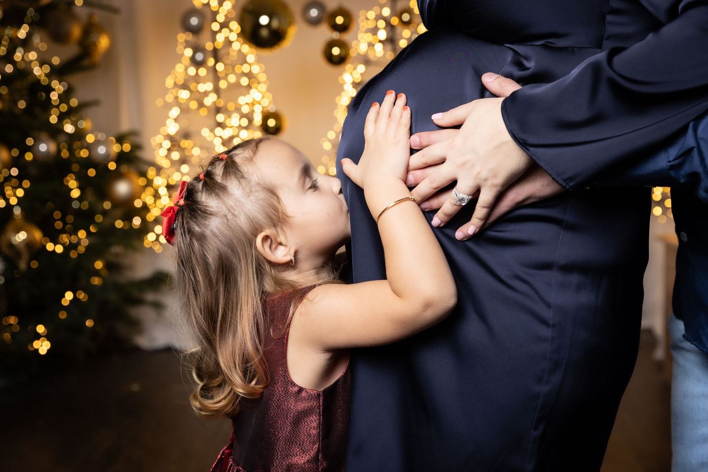 Kleines Mädchen küsst den Babybauch der Mama vor dem Weihnachtsbaum