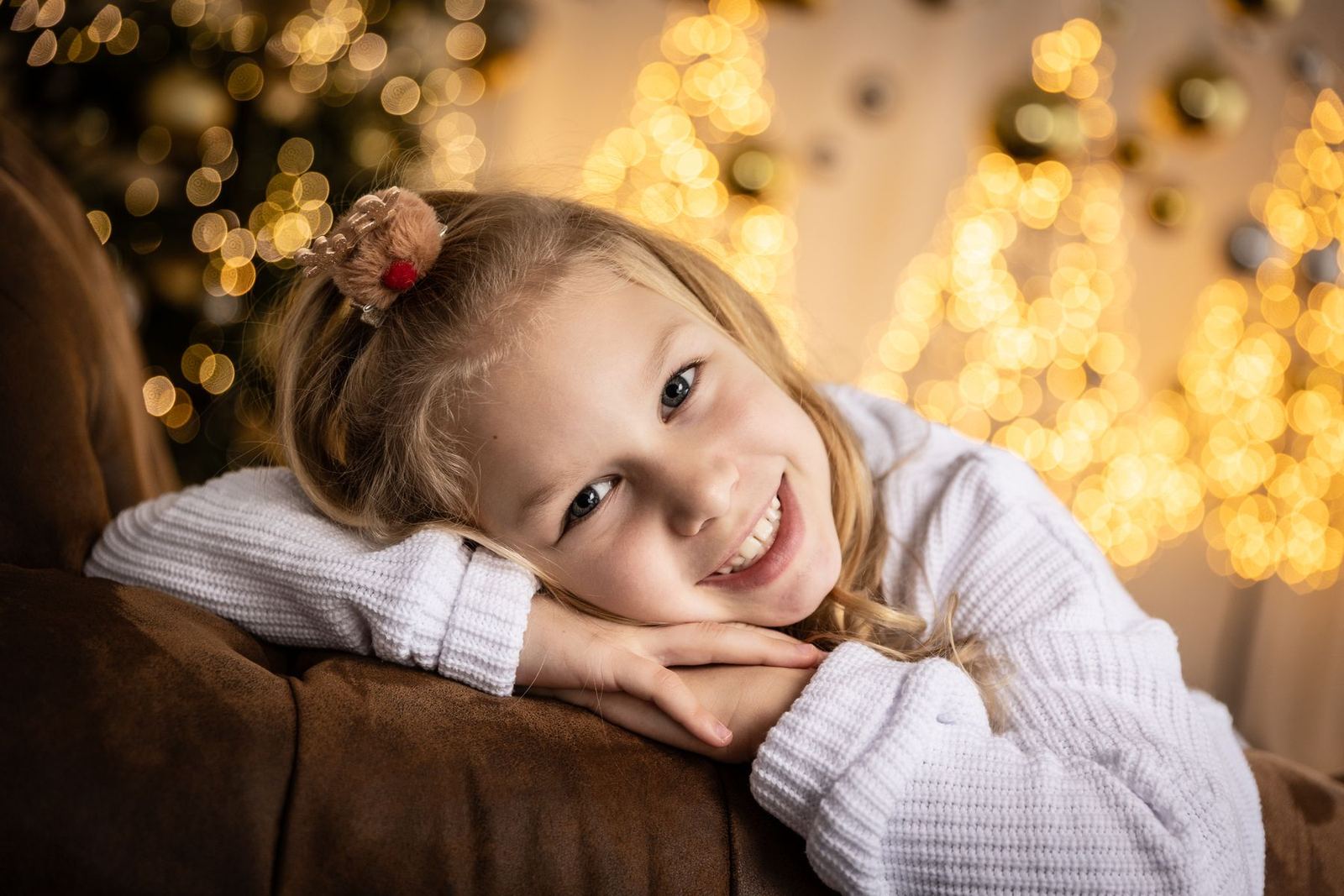 Mädchen im weißen Pullover liegt lächelnd auf dem Ledersofa vor warmen Weihnachtslichtern